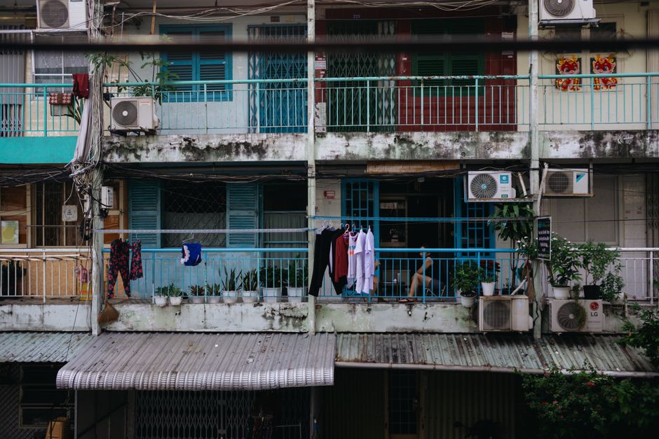 Weathered apartment balcony with clothes drying, plants, and air conditioners in an urban setting.