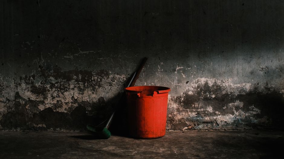A red bucket and broom leaning against a rough, textured wall in a dimly lit room.